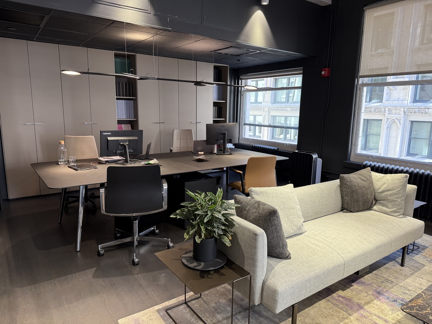 Blockarbeitsplatz mit verzweigter Pendelleuchte AVVENI PENDANT 6 Hinter einem grünen Sofa steht parallel eine Arbeitsplatztischgruppe mit Stühlen auf beiden Seiten, darüber ist eine schwarze Pendelleuchte, AVVENI PENDANT 6, abgehängt, die sich mehrfach verzweigt und an der runde flache Leuchtenköpfe jeweils das Ende der Verzweigung bildet.