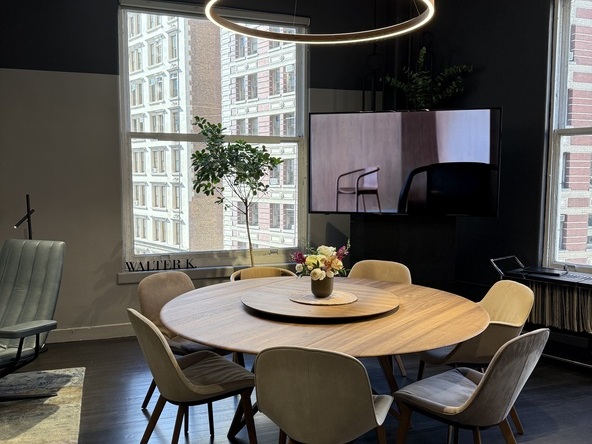 Essraum mit großen Fenstern und Blick auf den Broadway, in der Mitte ein runder Esstisch mit Stühlen aus Holz von Walter Knoll. Über dem Tisch hängt eine Ringleuchte von SATTLER der Serie DOPPIO, die den Tisch erleuchtet und einen runden Rahmen für die Essecke gestaltet.