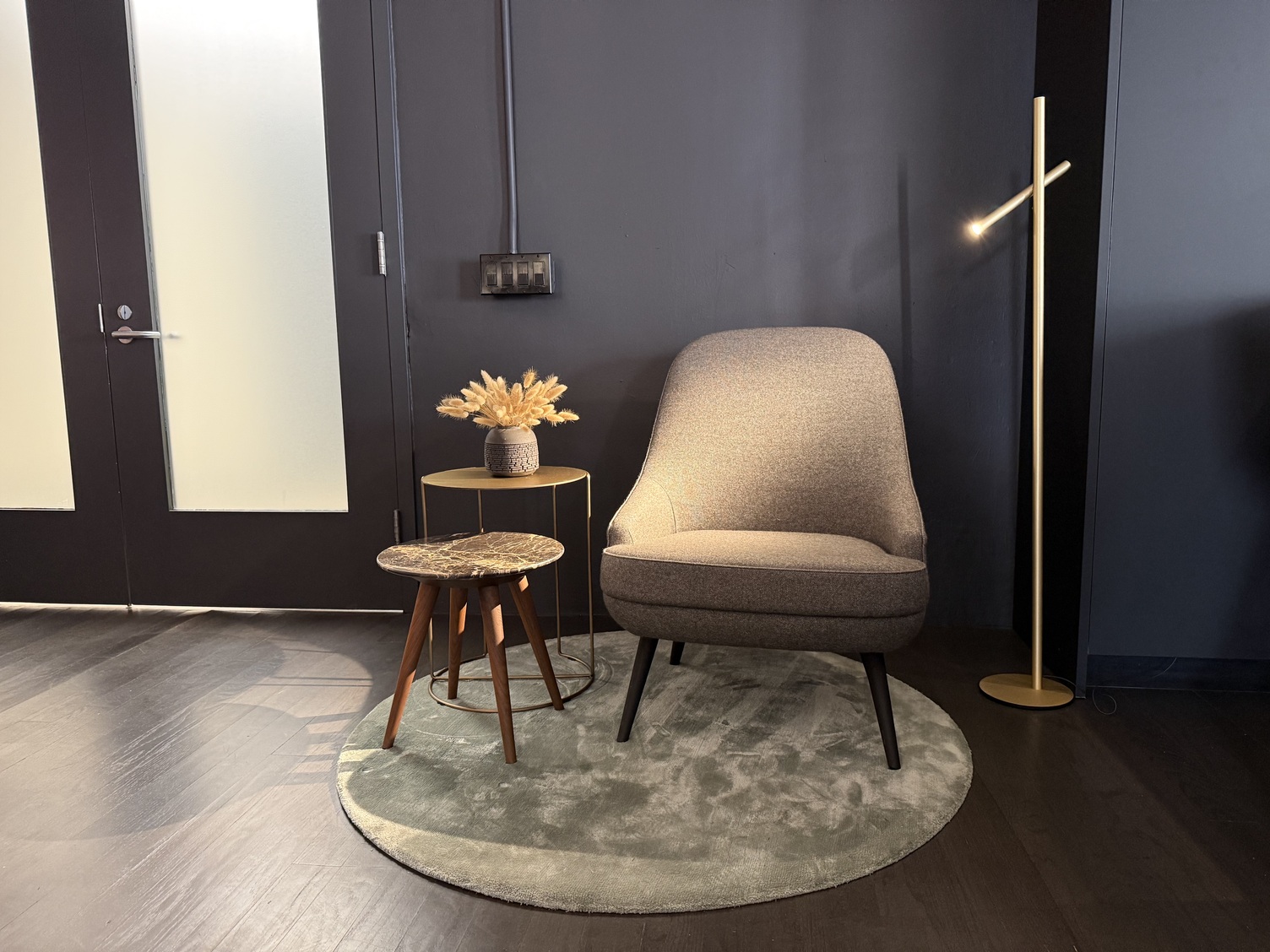 Leselicht mit Stehleuchte FINO FLOOR Platz neben einer Eingangstür. Graue Wand vor der ein hellgrüner runder Teppich liegt, auf dem steht ein gemütlicher heller Sessel und zwei kleine Beistelltische, rechts im Bild eine Stab-Stehleuchte, FINO PENDANT, mit einer Champagner-Gold-Farbenen Oberfläche, die die Sitzfläche beleuchtet.