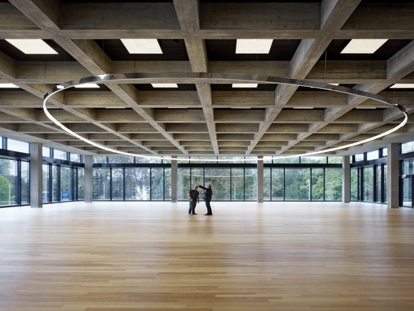 Elliptical pendant ring special luminaire (19m) free-form in the still large empty canteen of Roche AG, Building 67, Basel with two people standing underneath.  | © Ruedi Walti