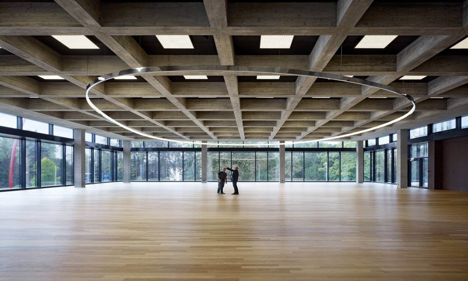 Elliptische Pendel-Ring-Sonderleuchte von SATTLER bei Roche | © Ruedi Walti Elliptische Pendel-Ring-Sonderleuchte (19m) Freiform in der noch großen leeren Kantine der Roche AG, Bau 67, Basel mit zwei Personen darunter stehend. | © Ruedi Walti