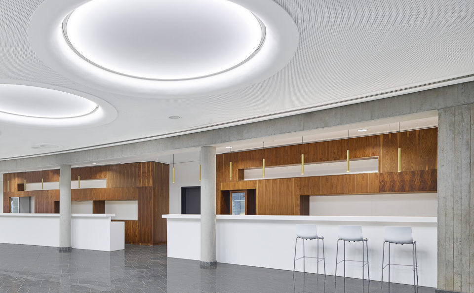 Großen Halle mit Steinboden und einer weißen Decke in der große rund Lichtfelder angeordnet sind, Blick geht in einen Thekenbereich, der als Nischen ausgeprägt ist und durch braune Holzwände hervorgehoben ist, zudem sind über die gesamte Theke in regelmäßigen Abständen goldene Stableuchten (TIAs) abgependelt.