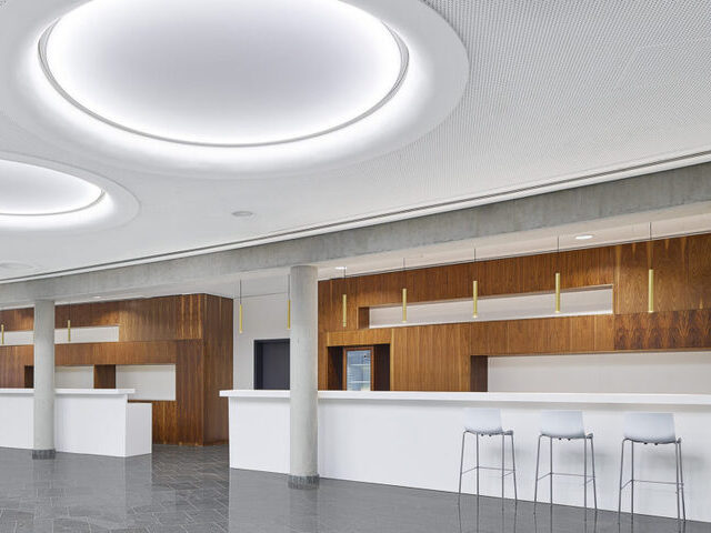 Großen Halle mit Steinboden und einer weißen Decke in der große rund Lichtfelder angeordnet sind, Blick geht in einen Thekenbereich, der als Nischen ausgeprägt ist und durch braune Holzwände hervorgehoben ist, zudem sind über die gesamte Theke in regelmäßigen Abständen goldene Stableuchten (TIAs) abgependelt. | © Zooey Braun